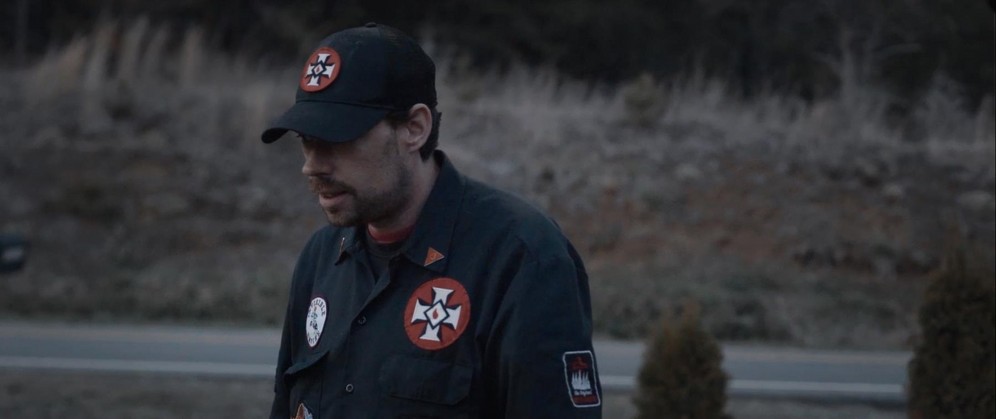 A man with a moustache is wearing a KKK uniform with a baseball cap and blue shirt with KKK badges sewn on. Still taken from RTD documentary Black Lives 8: Deadlock.