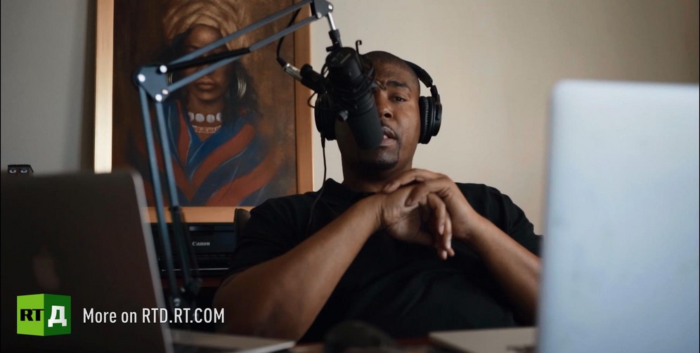 Tariq Nasheed is wearing headphones and speaking into a microphone. Still taken from RTD documentary Black Lives 8: Deadlock.