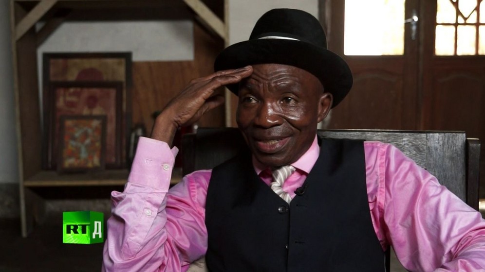 Severin Mouyengo, known as 'Le Salopard de la Sape', wearing a bowler hat, pink shirt and waistcoat. Screenshot taken from RTD documentary Congo Dandies.