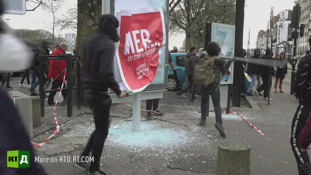 A person wearing a hoodie watches as another, also wearing a hoodie and a backpack, breaks the glass on urban furniture with an iron bar during a Yellow Vest protest in France.
Still taken from RTD 's documentary, Yellow Vest Fever.