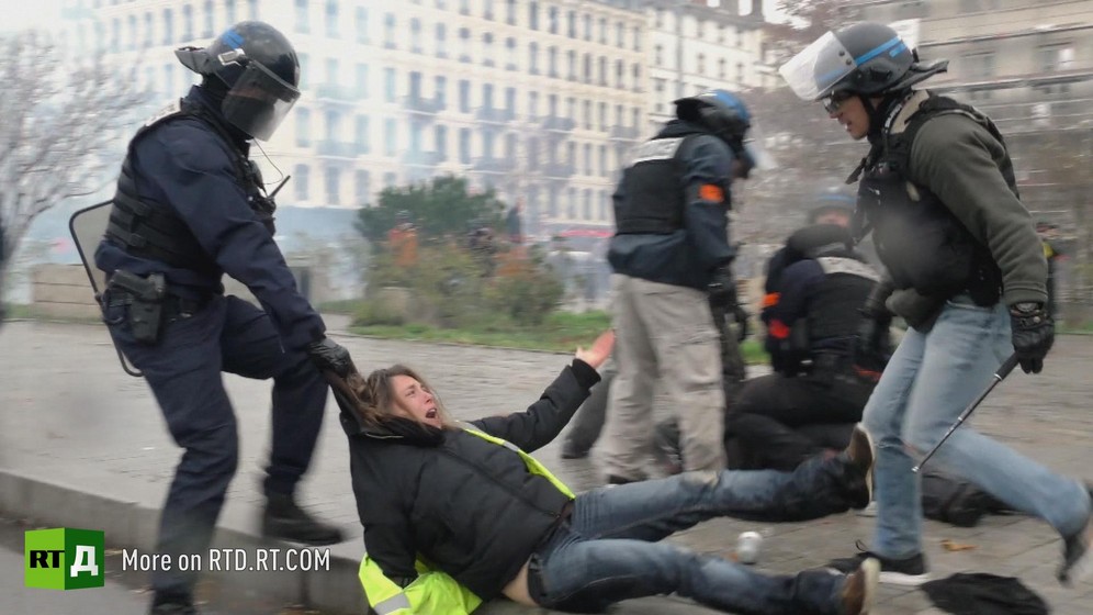 One riot policeman wearing a helmet, carrying a shield and a baton, drags a female Yellow Vest protester by the hood along the ground, while another policeman in plain clothes and wearing a helmet with the visor up towers over her, with other policemen neutralising another protester in the background.
Still taken from RTD 's documentary, Yellow Vest Fever.